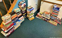 Photo showing multiple piles of stacked books on a carpeted floor near stairs and walls