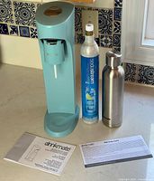 Photo showing the Arctic Blue Drinkmate OmniFizz soda machine, a Sodastream 60L CO2 cylinder, a stainless steel Drinkmate branded bottle, user manual, and warranty card on a kitchen counter.