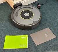 iRobot Roomba vacuum robot with charging dock and power cord, owner's guide, and warranty booklet on carpeted floor.