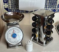 Photo showing the kitchen scale, rotating spice rack with spice jars, and sugar bowl on a kitchen counter against blue and white tiled backsplash.