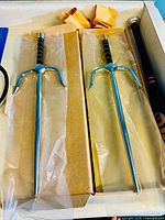 Pair of metal sais in packaging showing their metal construction, black wrapped handles, and pointed ends, stored in plastic sleeves within cardboard box.