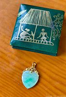 Green presentation box with gold-etched hut scene above jade heart pendant on wooden surface