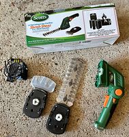 Photo of the full lot showing cordless trimmer device, two removable blade mounts, hedge blade, shear blade, charger, and original packaging box.