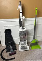 Front view of white Shark Navigator upright vacuum with black hose and accessory bag on left, green broom and dustpan on right, all on carpeted floor against cream wall.