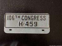 Photo of a metal license plate with raised black characters '106TH CONGRESS' and 'H-459' on a white background, two mounting holes at the bottom.