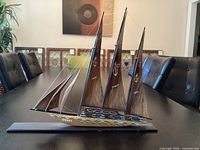 Full view of the wooden sailboat model showing its long base, three sails, and rigging, positioned on a table with background chairs.