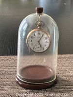 Antique Illinois pocket watch suspended inside a glass dome display case with wooden base and top finial, showing the watch face clearly