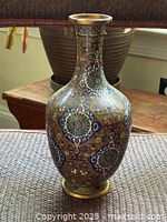 Photo showing full view of the vase with vivid cloisonne-style patterns, placed on a textured surface with a woven basket in background.