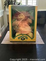Full view of the Cabbage Patch Kids doll in original box with yellow and green borders, visible through plastic window, seated wearing pink dress.