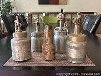 Top-down view of five vintage metal paint sprayers and one vintage metal oil can, showing varying sizes and designs on a dining table.