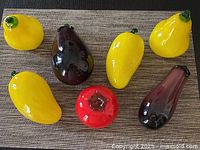 Top view of seven glass decorative fruits on a woven placemat, showing four yellow mango-shaped fruits, two dark purple eggplants, and one red pomegranate.