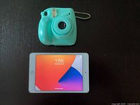 Front view of Fujifilm Instax Mini 7+ instant camera in turquoise and Apple iPad Mini powered on with colorful home screen.