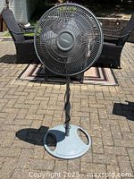 Photo of a Laslo floor fan placed on a stone patio with outdoor furniture in background, showing fan head, adjustable pole, and round base.