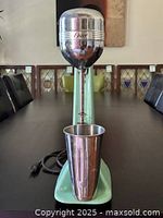 Full view of light green Oster stand mixer with aluminum mixing cup attached, showing power cord and base.