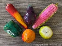 Photo showing six colorful paper mache fruits including carrot, eggplant, ear of corn, bell pepper, orange, and apple.