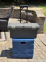 Closed Rubbermaid black toolbox on top of Staples blue storage box with handle extended, outdoor patio background
