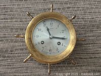 Front view of Schatz ship bell clock mounted in brass ship wheel frame showing clock face and hands.