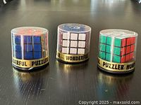 Three vintage 3x3 Rubik Cubes housed in original clear plastic cases with a gold 'PUZZLER WONDERFUL' band.