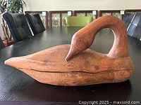 Photo shows wooden duck decoy on a dark table surface, carved with stylized details showing the duck's head bent backward.
