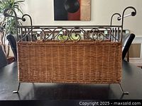 Front view of the wicker and metal magazine rack showing woven wicker panels set inside an ornamental metal frame with curved scrollwork on top.