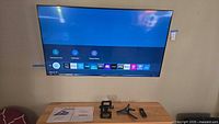 Samsung Smart TV mounted on wall displaying home screen with apps. Below TV on table: two wall mounts (one fixed, one swivel), TV remote, and TV legs, along with user manual booklets.