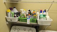 Shows assorted household cleaning bottles and products arranged on a wire laundry room shelf and on top of washing machine and dryer