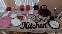 Full top-down view of assorted kitchen items on a beige tablecloth including wall decor, ceramics, linens, kettle set, clock, black wire basket, and other accessories.