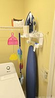 Black & Decker steam iron seated in mount on wall above blue ironing board along with hanging dusters and pink dustpan/fly swatter.