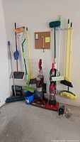 Wide view showing cleaning tools including brooms, mop, bucket, Dustbuster, and two upright Dirt Devil vacuums against a wall in garage.