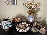 Full view of all items in the lot including faux flower arrangement in painted vase, wicker bowl with decorative balls, angel girl statues, glass hanging angel and small decorative jars.