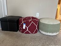 Three ottomans side by side on carpeted floor: black leather ottoman with storage, burgundy patterned fabric ottoman, green woven fabric ottoman