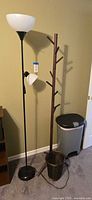 View of black metal floor lamp with large upward white shade and smaller reading lamp below, wooden coat tree with branches for hanging, flip top trash can, and small round wicker wastebasket all situated in corner of room.