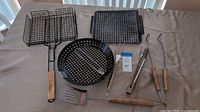 Photo showing a set of assorted grilling accessories laid out on a table, including two rectangular vegetable baskets with wooden handles, a round perforated grill pan, a square perforated grill tray, metal meat fork, two grilling tongs, and a spatula, all with wooden handles.