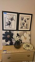 Two 18x22 framed black-and-white floral prints on the wall above two 18x18 floral canvas prints on the dresser, faux floral arrangement in a decorative dark glass vase, faux floral wreath, and small wall decor piece on dresser.