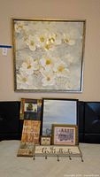 Framed floral wall art with white and yellow flower blossoms, along with multiple smaller decorative signs and plaques arranged below on black chairs.
