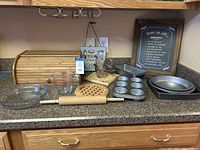 Wooden bread box, metal muffin pans, loaf pans, glass measuring cups, rolling pin, trivet, kitchen wall art, and cookie roller on kitchen countertop.
