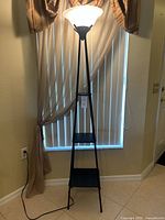 Full view of the black metal floor lamp with frosted white conical lampshade and three square shelves.