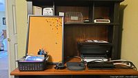 Wooden desk with cork bulletin board, pen holder, container with paper, and HP Deskjet 3050 printer on right side with cords and power strip below