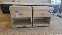 Pair of matching light wood nightstands on carpet in front of a mattress base, showing overall front and side view.