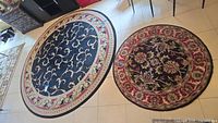 Two round area rugs side by side on tile floor, larger rug 95" diameter with black background and beige-red border, smaller rug 48" diameter with black center and red floral border.