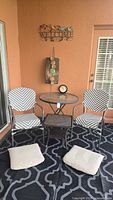 Full view of patio set including round glass top table, two chevron pattern chairs, two seat cushions on floor, decorative wall art, and small outdoor clock on table.
