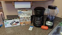 Photo showing Mr. Coffee 12-cup coffeemaker, Hamilton Beach blender, Sharper Image mandoline slicer box, manual ricer/masher box, air fryer accessories box, and kitchen Mama can opener on kitchen counter.