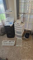Wide view of all Sterilite plastic drawer units and assorted organizers arranged on carpet near window with blinds.
