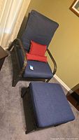 Wicker lounge chair with high back blue cushion, red accent pillows, and matching ottoman with blue cushion on carpeted floor.