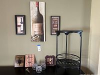 Wide view of entire wine decor set including three wall art pieces, wine glasses with text, wine stopper, and black metal side table.
