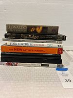 Stack of eight books showing titles: Leonardo, Stage Makeup Ninth Edition, Men's Garments 1830-1900, Folk Costumes of the World, The New Artist's Manual, Costume Design Screencraft, and two unidentified books under.