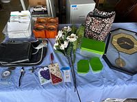 Overview of assorted home items on table including greeting cards, orange candle holders, black leather bag, cheetah print lunchbox, green containers, artificial flowers, bookmarks, and a wall clock
