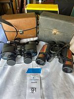Two pairs of Bushnell binoculars displayed with two carrying cases, one brown, one gray, and the box behind.