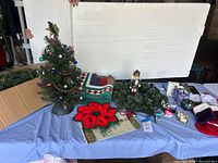 Photo shows a blue table covered with various Christmas decorations including a decorated mini Christmas tree, a wooden nutcracker figure, a green wreath with a large red poinsettia, several holiday stockings, a folded quilt with Christmas patterns, and small Christmas figurines and collectibles.