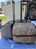 Photo showing three rolling crafting bags side by side with telescoping handles extended, displaying different fabric patterns including gray floral, beige paisley, and dark floral.